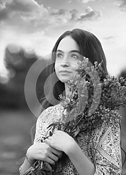 Black and white portrait with a young girl looking into the distance, holding a bouquet of lilac lupins in her hands. Stunning sky