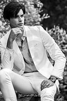 Black-white portrait of young handsome fashionable man in white suit against nature background