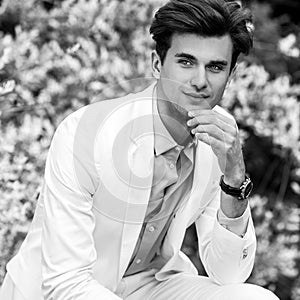 Black-white portrait of young handsome fashionable man in white suit against nature background