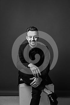 Black and white portrait of a young handsome man in a suit sitting on box, smiling and looking at the camera