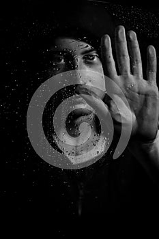 Black and white portrait of a young hooded man holding his hand on a glass covered in water drops - focus on the glass