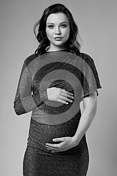 Black and white portrait of young pregnant female in grey sequin dress with hands near pregnant belly