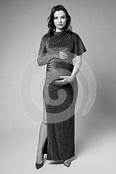 Black and white portrait of young pregnant female in grey sequin dress with hands near pregnant belly