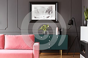 Black and white poster hanging on molding wall in dark living room interior with green cupboard with plant and vases, metal lamp