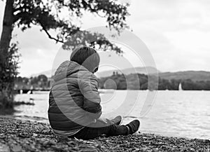 Black and white rear view of lonely kid head down and looking at his feet, Sad child sitting alone by te lake, Bored little boy
