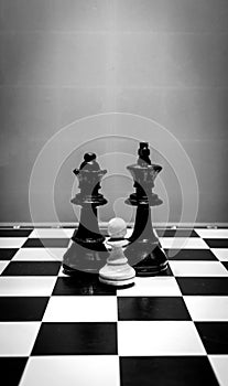 Black and white shot of chess pieces of a black king and queen and a white pawn standing in front of them on a chessboard