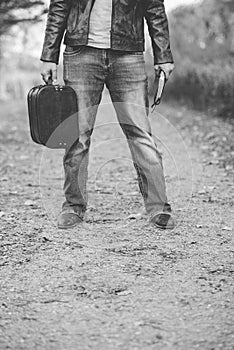 Black and white shot of a male standing on an empty pathway while holding the bible and old suitcase