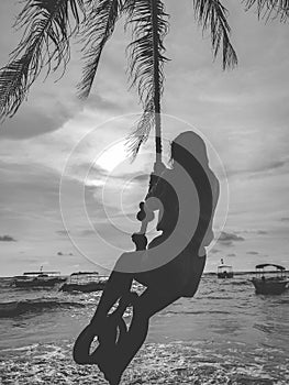 Black and white silhouette of young woman swinging on the swing on palm at ocean beach