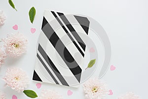 black and white striped notebook, pink plastic hearts and pink flowers of chrysanthemums and green leaves on a white table.