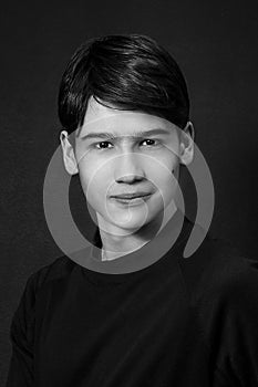 Black and white studio portrait of a brown-eyed, dark-haired, smiling teenager with a mole on his cheek in black clothes on a