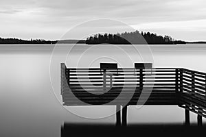 Black and white tranquil scenery of a lake with pier and two chairs