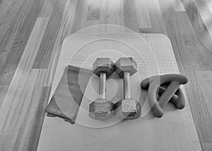 Black and white version of Overhead view of a pair of dumbbells, theraband exercise bands, and a yoga mat on hardwood floor