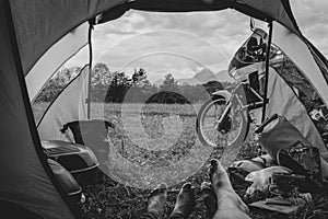 Black and white view from camp tent door. Motorcycle adventure bike on the background of green grass, forest and mountains under
