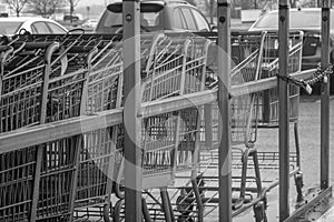 Black and white view of group of shopping carts in a metal enclosure in a parking lot with cars in the background.