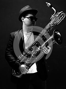 Black and White. Young Saxophonist in a Black Jacket, Hat and Sunglasses Holds a Baritone Saxophone. Dark Background