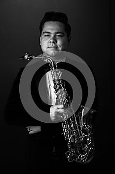 Black and White. Young Saxophonist in a Black Jacket, Hat and Sunglasses Holds a Baritone Saxophone. Dark Background