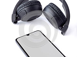 Black wireless headphone and mobile smartphone with a blank screen mockup lay on the surface of a white background