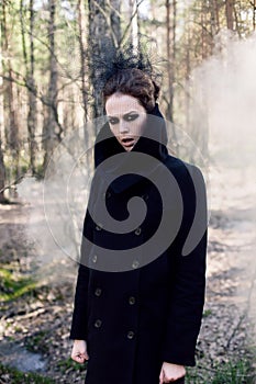 black witch in long coat and black eyes stands in an autumn gloomy forest with fog for halloween