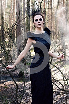 black witch in long dress and black eyes stands in an autumn gloomy forest with fog for halloween