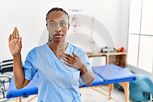Black woman with braids working at pain recovery clinic swearing with hand on chest and open palm, making a loyalty promise oath