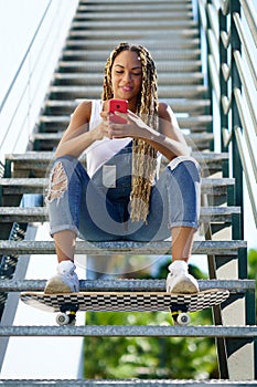 Black woman with coloured braids, consulting her smartphone with her feet resting on a skateboard.