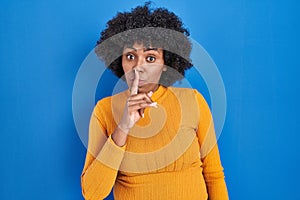 Black woman with curly hair standing over blue background asking to be quiet with finger on lips