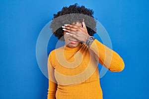 Black woman with curly hair standing over blue background covering eyes with hand, looking serious and sad