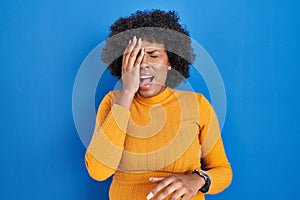 Black woman with curly hair standing over blue background yawning tired covering half face, eye and mouth with hand
