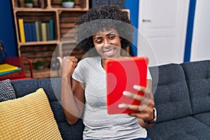 Black woman with curly hair using touchpad sitting on the sofa pointing thumb up to the side smiling happy with open mouth