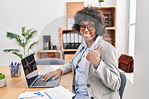 Black woman with curly hair wearing call center agent headset at the office doing happy thumbs up gesture with hand