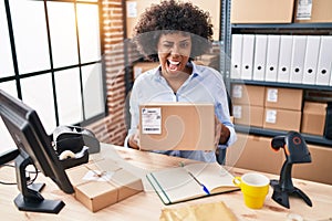 Black woman with curly hair working at small business ecommerce holding box winking looking at the camera with sexy expression,