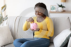 Black woman putting coin in piggy bank, sitting on couch at home