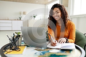 Black woman sitting at desk, using pc writing in notebook
