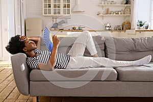 Black woman suffering from heatstroke flat without air-conditioner waving fan lying on couch at home