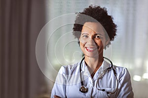 Black woman in white lab coat with stethoscope standing in medical office for exam room at clinic