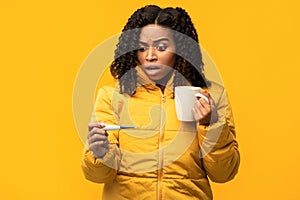 Black Woman In Winter Jacket Holding Thermometer On Yellow Background