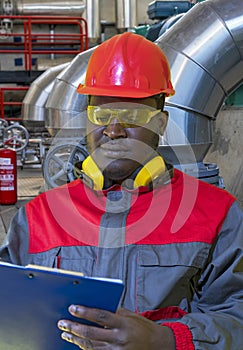 Black Worker In Protective Workwear Writing On Clipboard In Industrial Interior