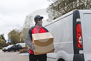 Black young adult delivery guy wearing work uniform and black cap smiling walking by white van holding carboard box