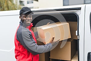 Black young adult delivery guy in work uniform and black cap unloading packages from white van looking into camera