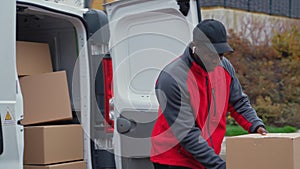 Black young adult delivery guy in work uniform and black cap unloading packages from white van looking into camera