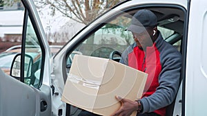 Black young adult delivery guy in work uniform sitting in door of white van looking at cardboard box parcel.