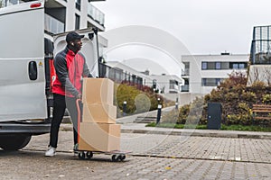 Black young adult delivery guy in work uniform unloading cardboard box parcels from white van onto trolley. Horizontal