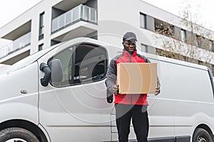 Black young adult delivery man wearing work uniform standing outside of white van holding cardboard box parcel