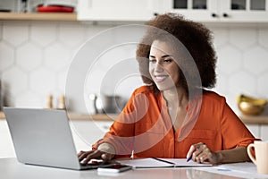 Black Young Freelancer Woman Working With Laptop In Kitchen And Taking Notes
