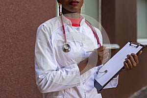 Black young woman doctor, in a white coat, with a phonendoscope, holds a folder for medical papers in a rue, against the