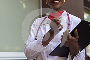 Black young woman doctor, in a white coat, with a phonendoscope, holds a folder for medical papers in a rue, against the