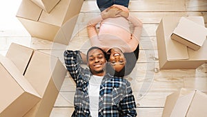 Black young woman with her handsome husband lying among cardboard boxes on floor of their new home, top view