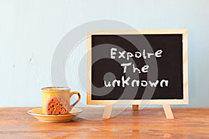 Blackboard with the phrase explore the unknown next to cup of coffee and cookie
