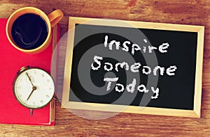 Blackboard with the phrase inspire someone today written on it. over wooden table with coffee and vintage clock