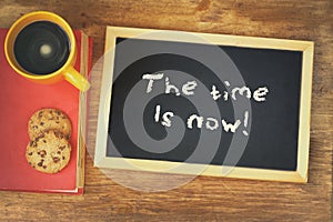 Blackboard with the phrase the time is now written on it, next to coffee cup and cookies